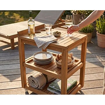 Luxe teakhouten serveerwagen op een zonnig terras, gestyled als buitenbar met wijn en hapjes. Een hand pakt een wijnglas van het bovenblad.